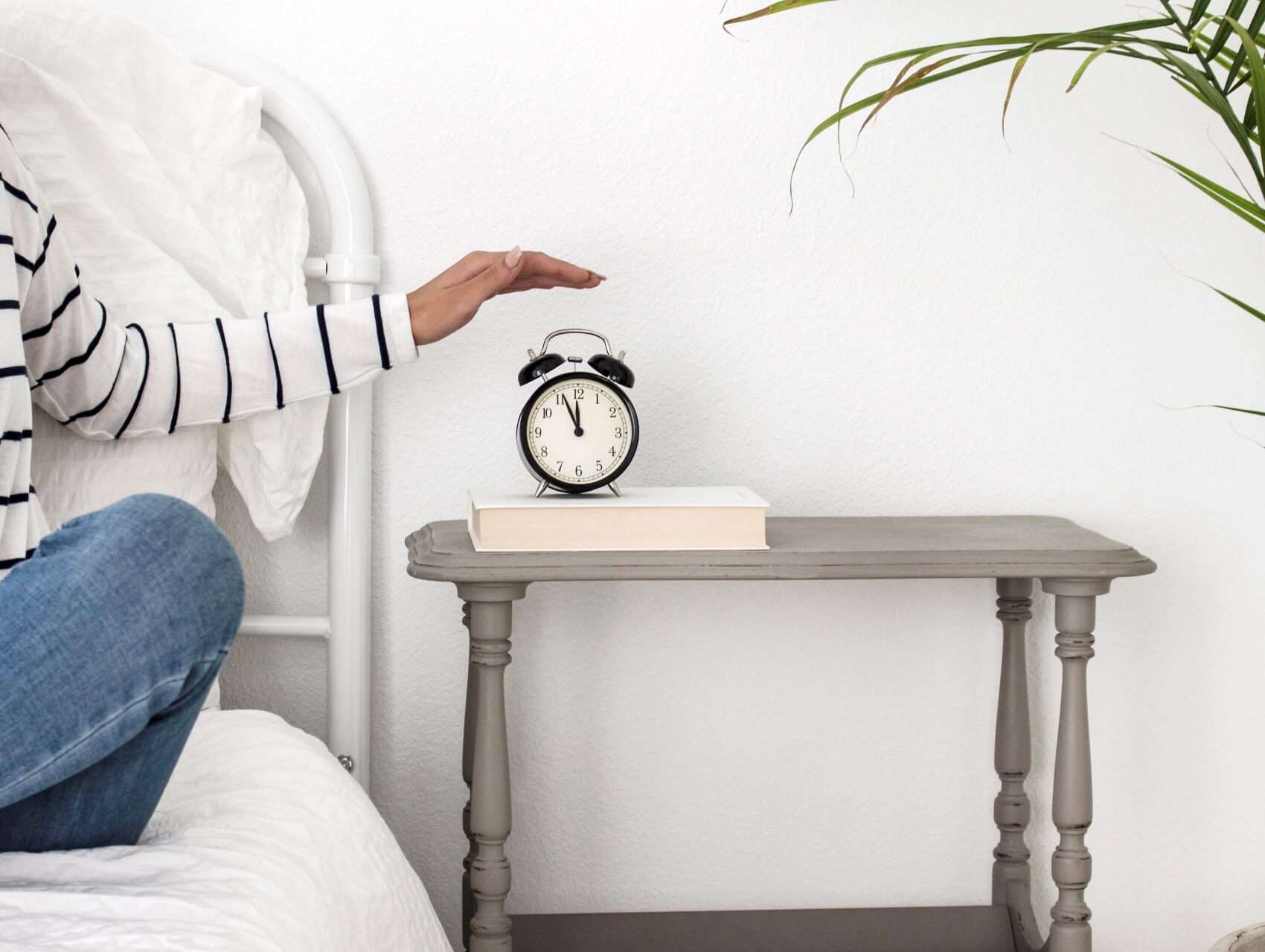 woman sitting on her bed with her hand on the alarm clock that is sitting on a sidetable - morning routine for bloggers